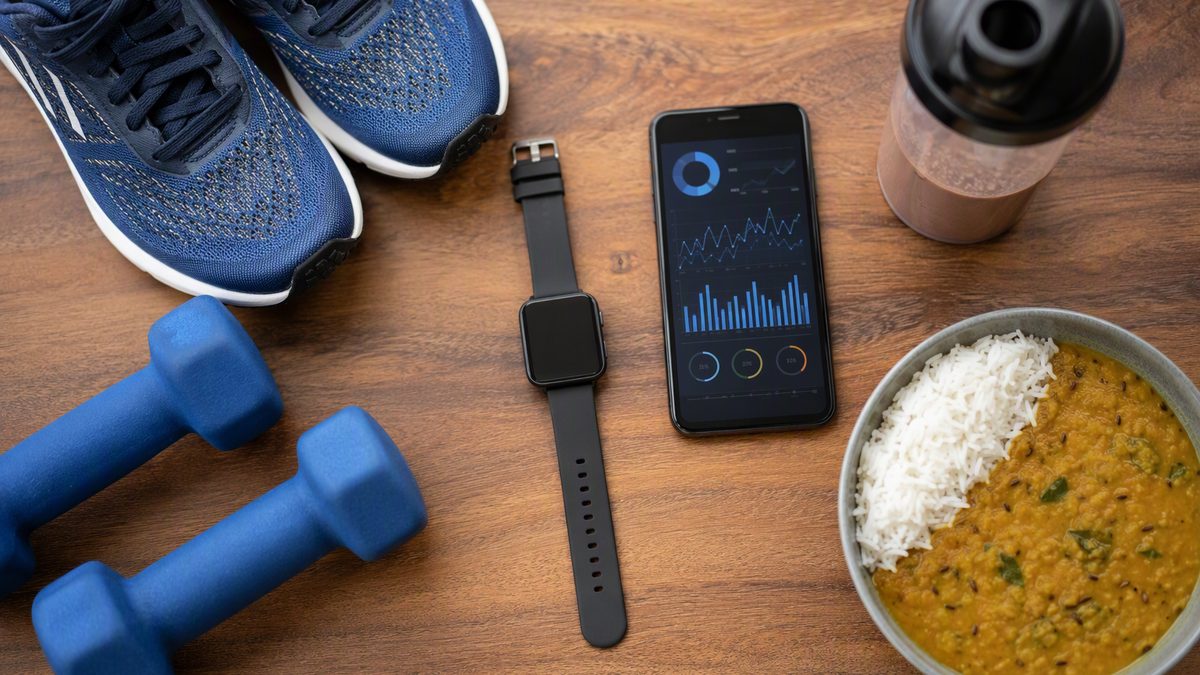 Flat-lay of fitness life: running shoes, dumbbells, a smartwatch, a phone with charts, a shaker and a bowl of dal and rice on a wooden table